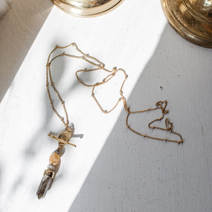 Labradorite lariat necklace coiled on a sunlit white surface beside brass bowls