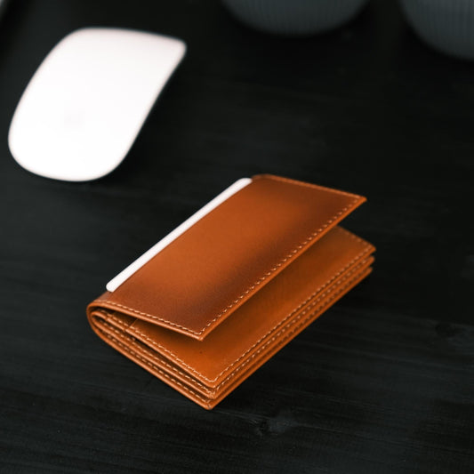 Tan leather card holder lying closed on a dark desk beside a computer mouse, a card edge showing