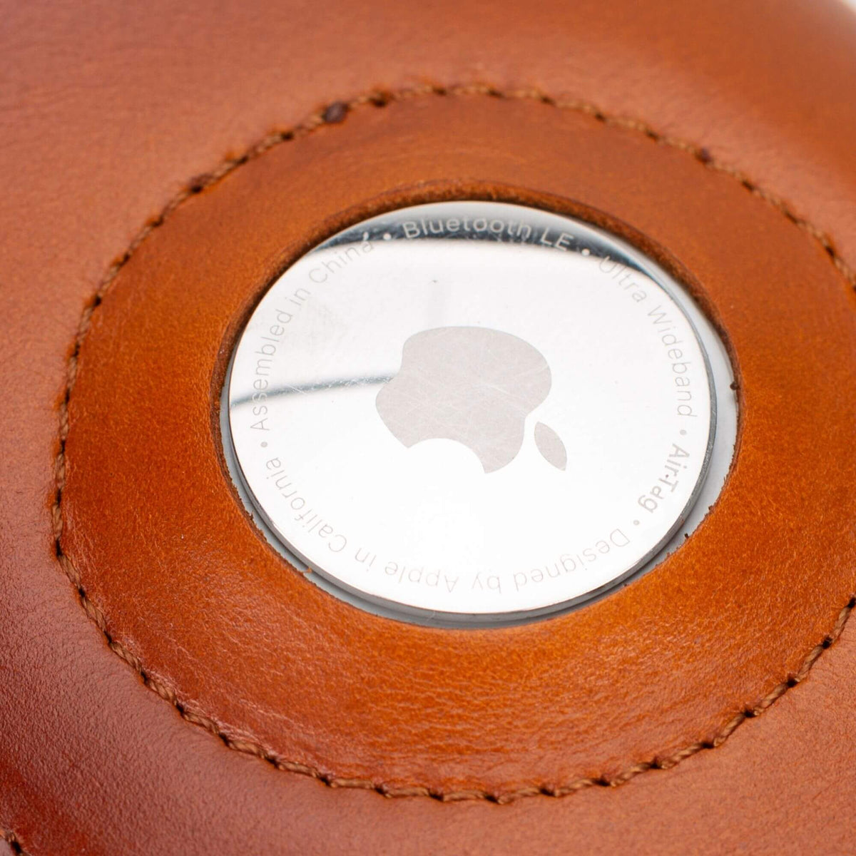 Close-up of an AirTag seated in the round window of the tan leather wallet, stitching visible