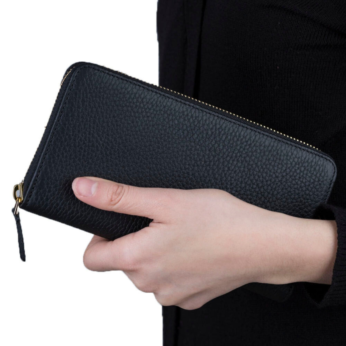 Women's zip around wallet in black pebbled leather held in one hand, the gold zip pull hanging