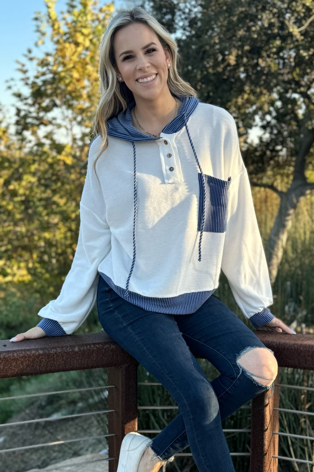 White knit hoodie with a blue pocket, model seated on a railing