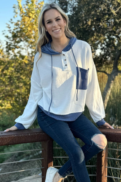 White knit hoodie with a blue pocket, model seated on a railing